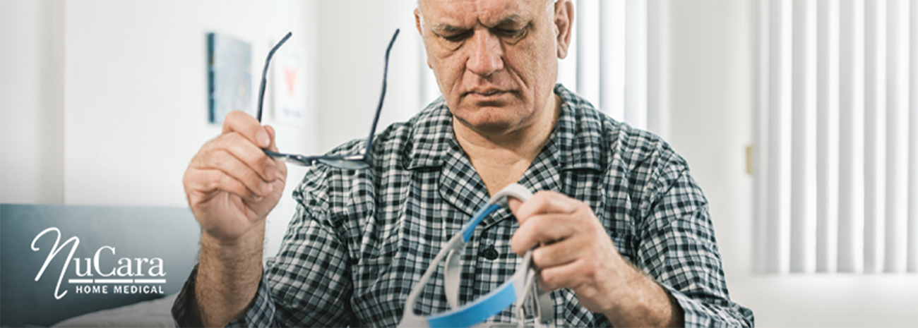 Man sitting on bed confused with glasses in one hand and CPAP mask strap in the other