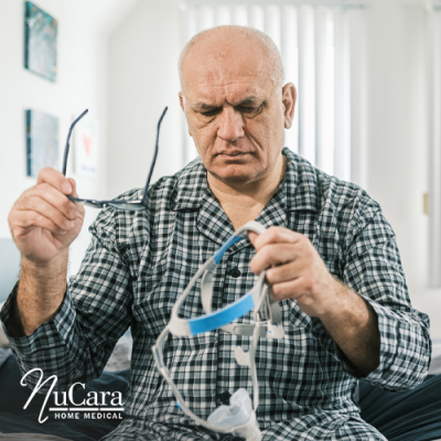 Man sitting on bed confused with glasses in one hand and CPAP mask strap in the other